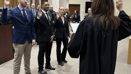 A white male, black male and white female stand with right hands raised in a court room looking at a female judge who also has right hand raised