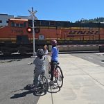 two children on bikes watching train as it travels through a street crossing