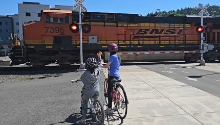 two children on bikes watching train as it travels through a street crossing