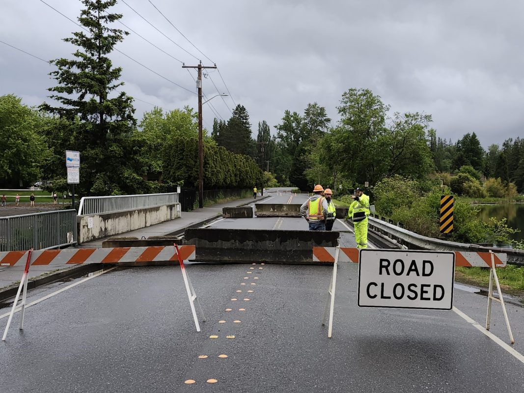 road closed sign posted in roadway near bridge with several construction workers in high visibility clothing in the background talking