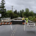 road closed sign posted in roadway near bridge with several construction workers in high visibility clothing in the background talking