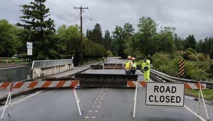 road closed sign posted in roadway near bridge with several construction workers in high visibility clothing in the background talking