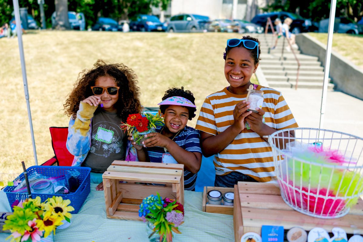 Three children smiling as they sell handmade crafts at their booth