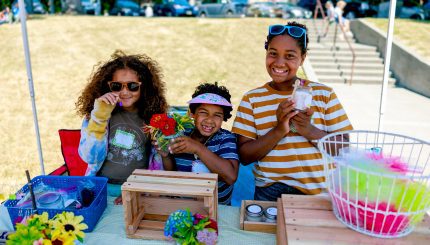 Three children smiling as they sell handmade crafts at their booth