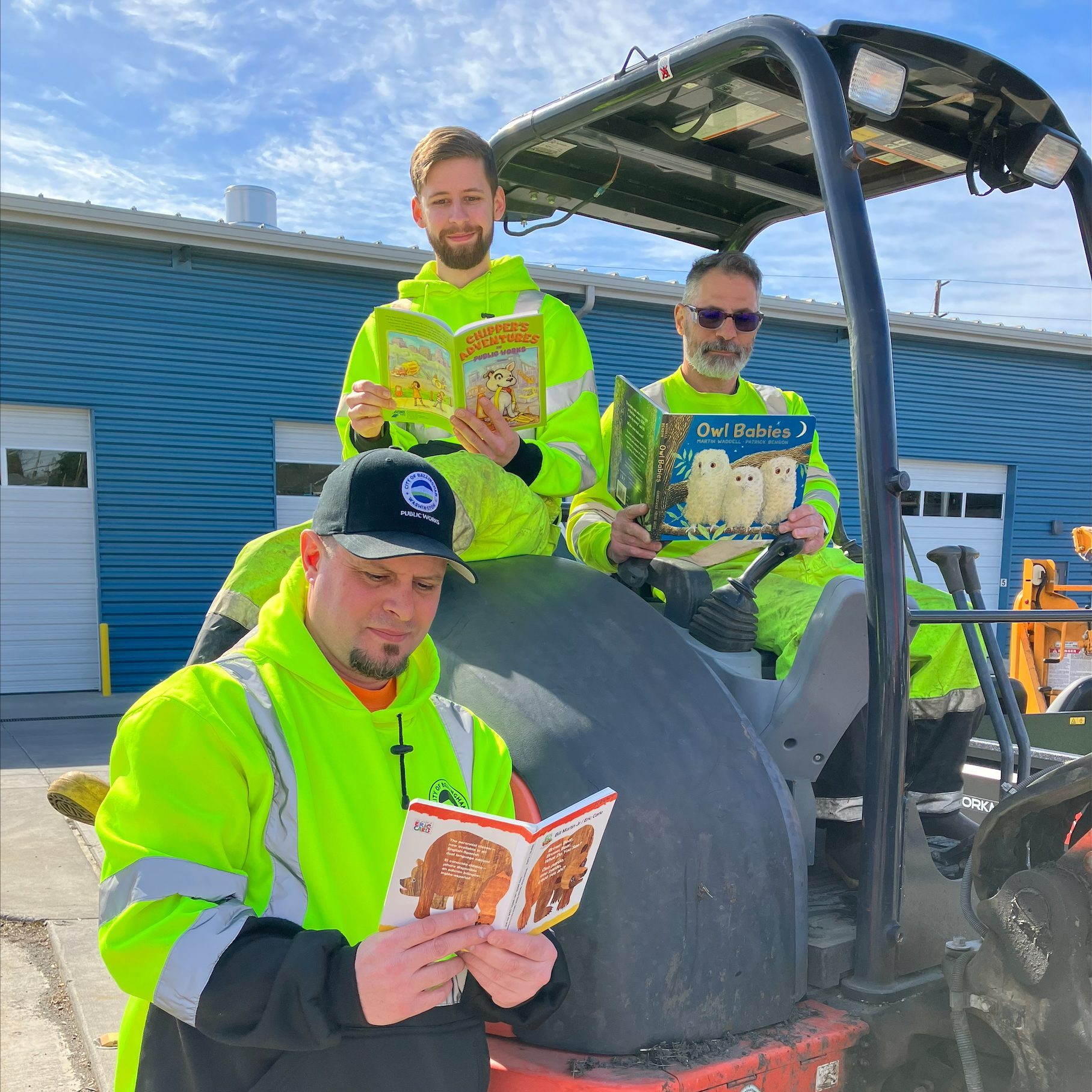 Three public works crew members reading children's books on a piece of machinery