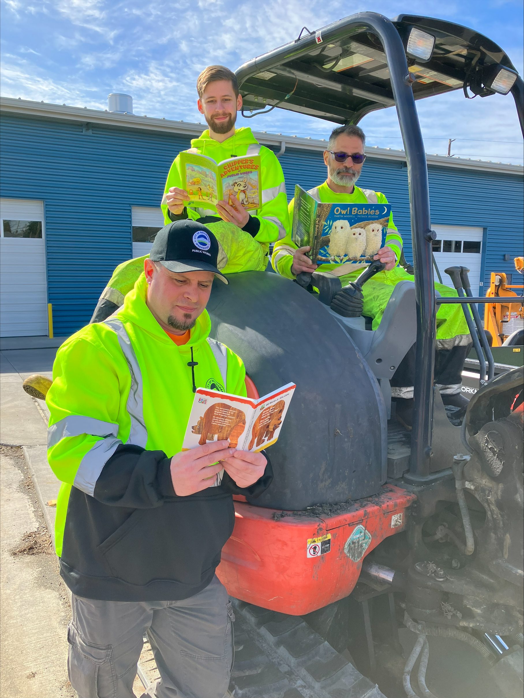 Three public works crew members reading children's books on a piece of machinery