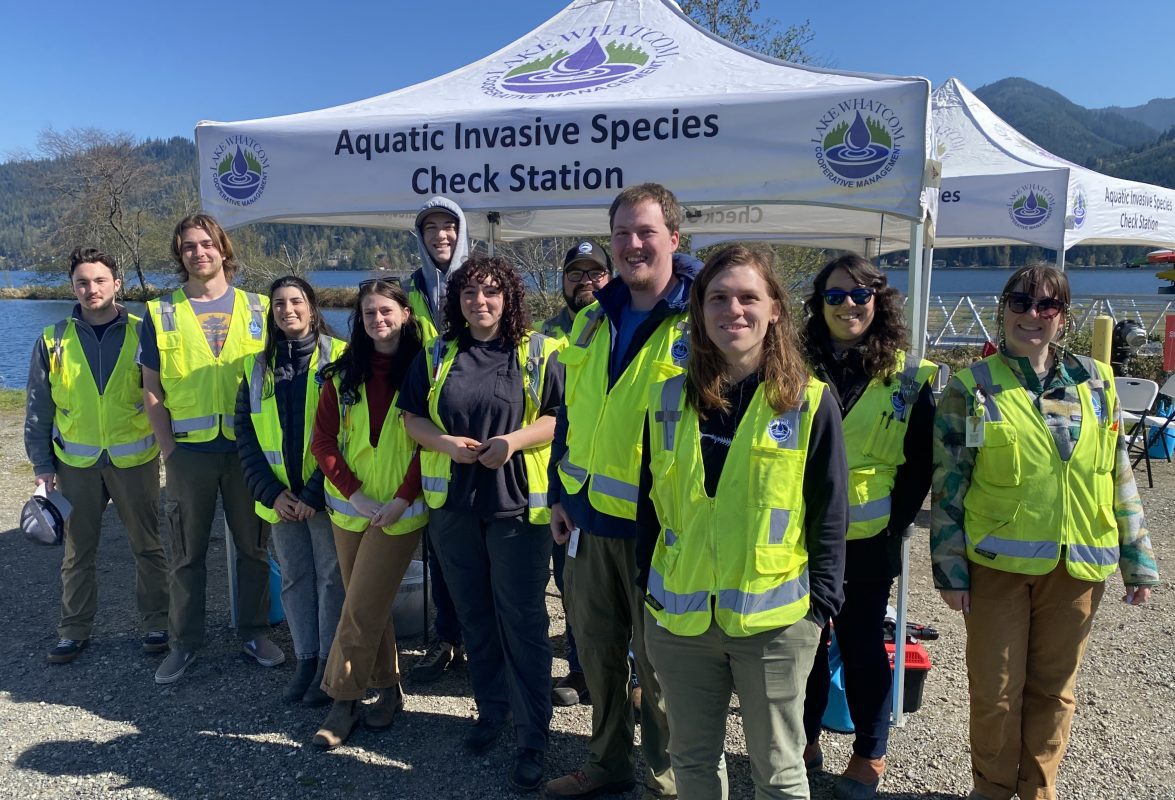 12 adults with reflective yellow vests stand in front of a white canopy that says Aquatic Invasive Species Inspection Station