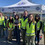 12 adults with reflective yellow vests stand in front of a white canopy that says Aquatic Invasive Species Inspection Station