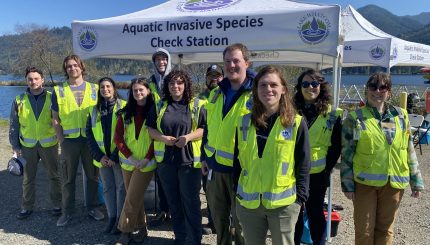 12 adults with reflective yellow vests stand in front of a white canopy that says Aquatic Invasive Species Inspection Station