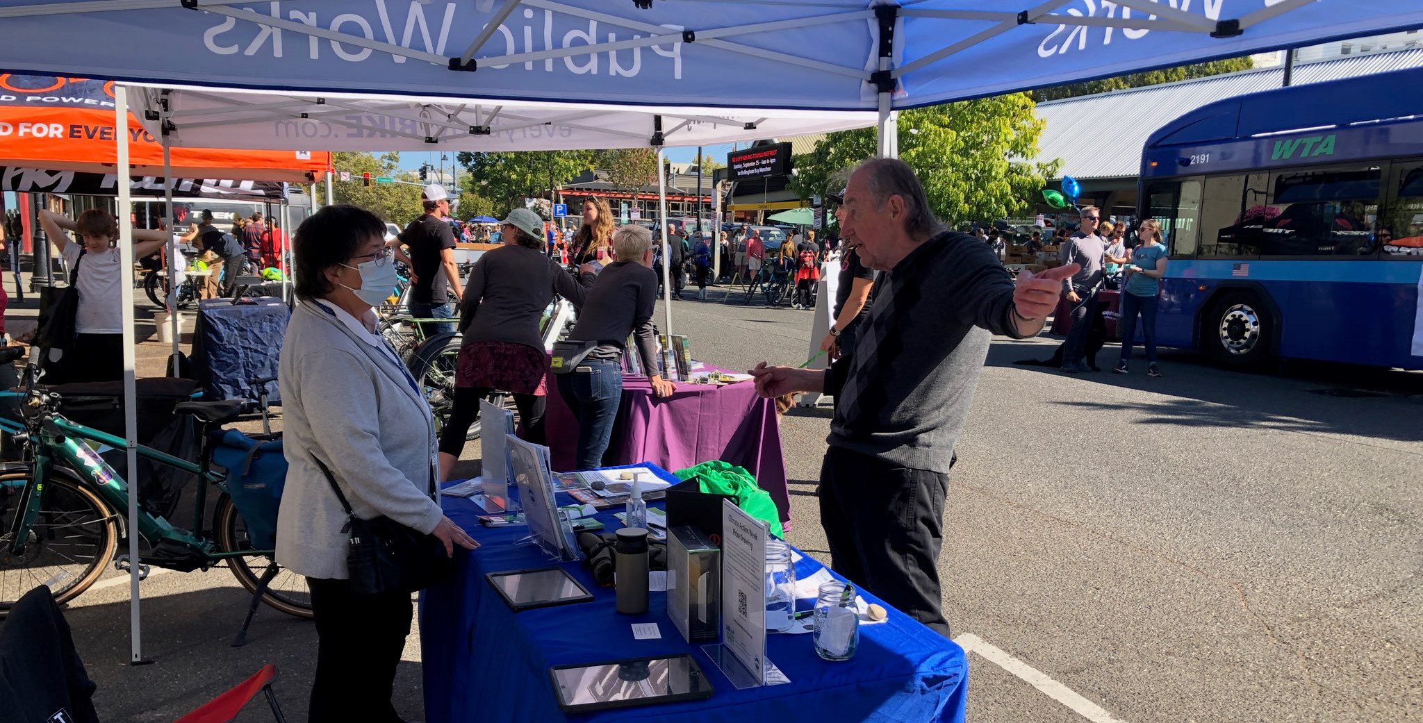 Two individuals talking at an informational table outside. People are in the background walking around the Farmers Market. There is a parked WTA bus and several bikes in the background.