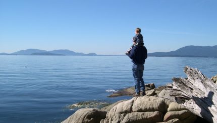Adult with child on their shoulders standing on a rock looking out into Bellingham Bay.