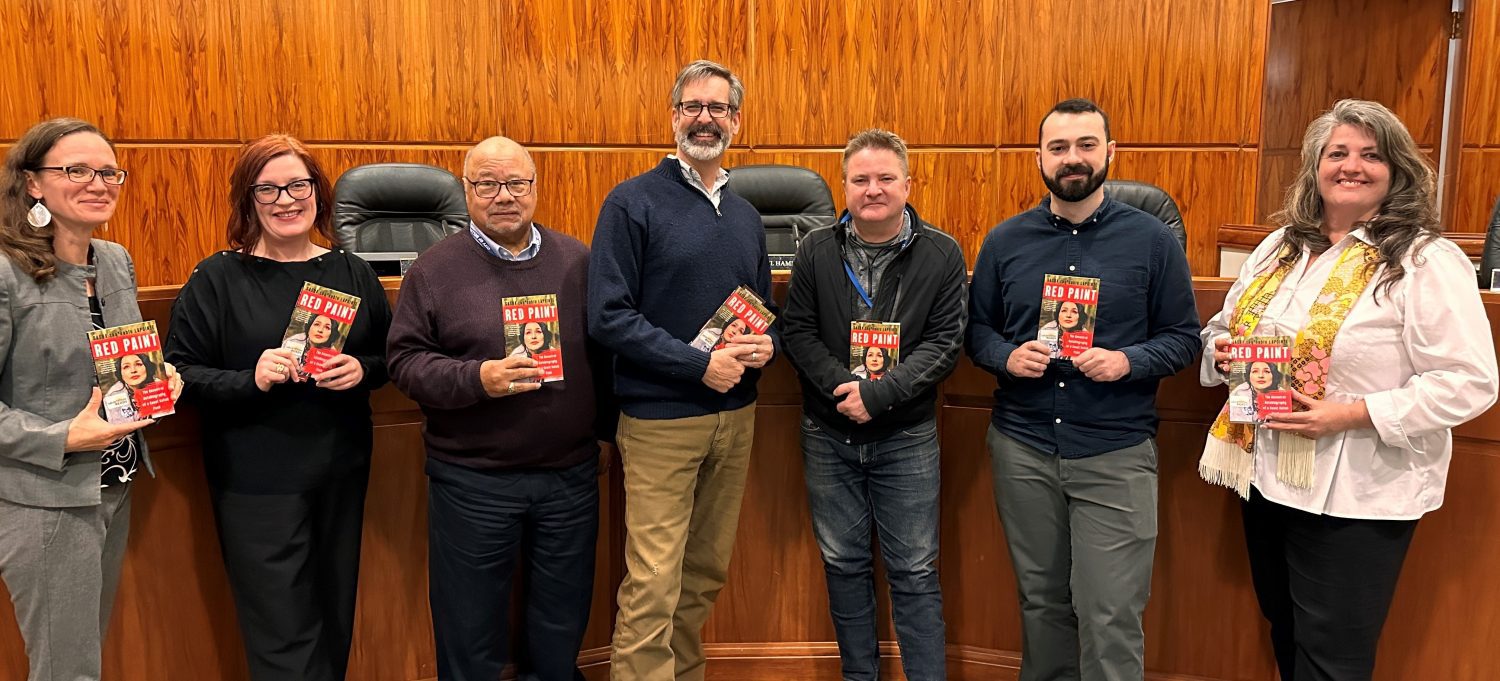 City Councilmembers pose in City Council Chambers, each holding a copy of Red Paint.