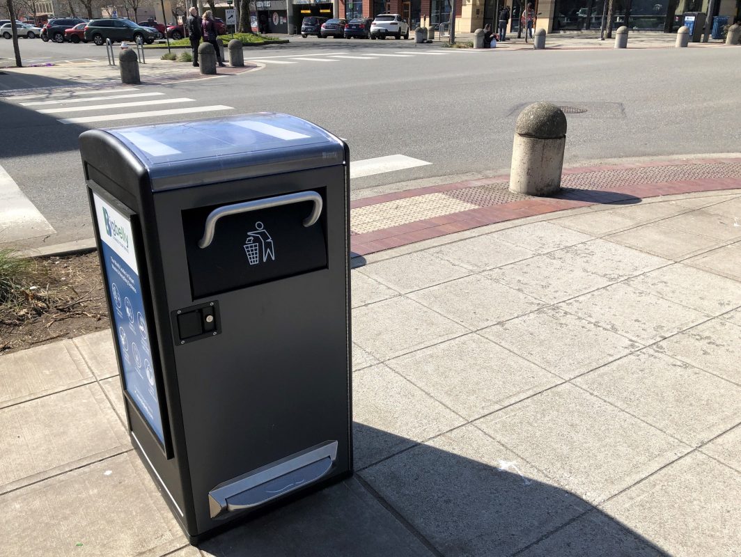 Covered garbage bin on sidewalk at intersection. 