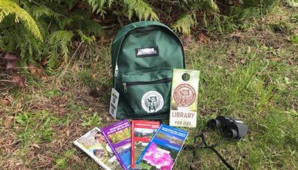 Green backpack sitting on grass with a Discover pass, park maps, and binoculars next to it.