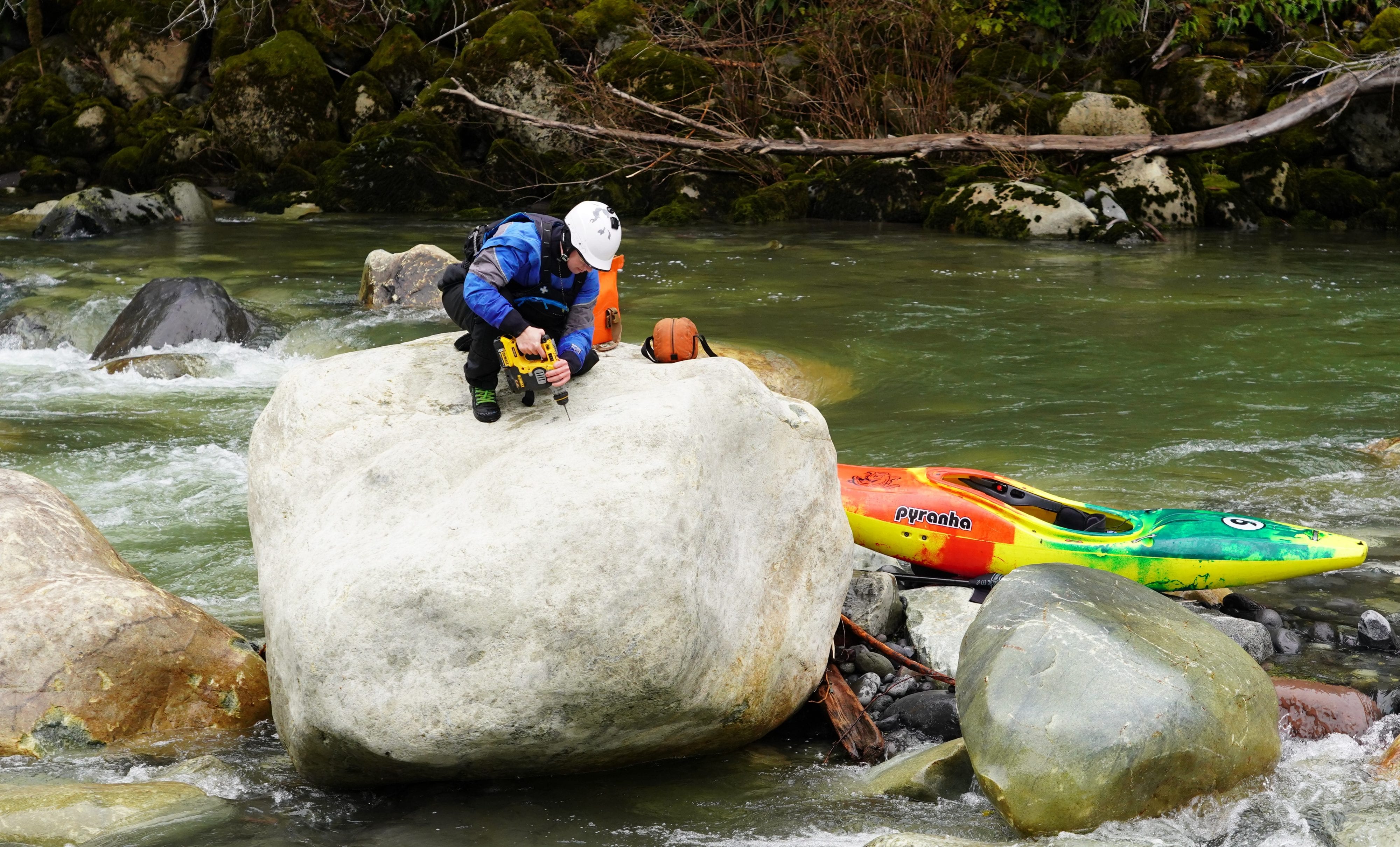Kayaker sitting on top of a boulder in the middle of the river, drilling a hole into the boulder