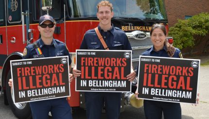 Three firefighters standing in front of a firetruck holding yard signs that read "Fireworks Illegal In Bellingham, subject to fine. Bellingham Municipal Code 10.24.130"