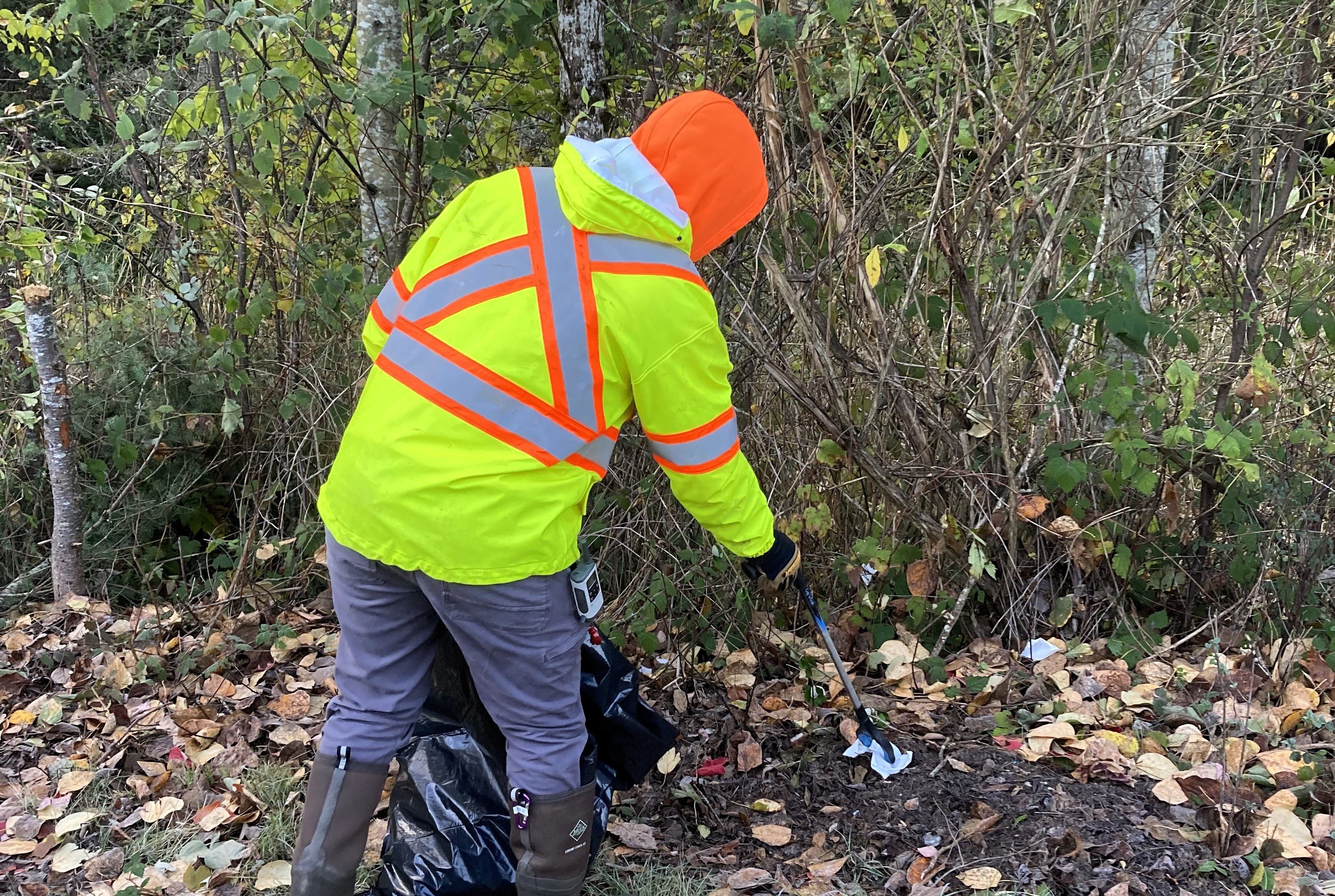 Person in brightly colored safety jacket with orange hood using litter grabber to pick up litter