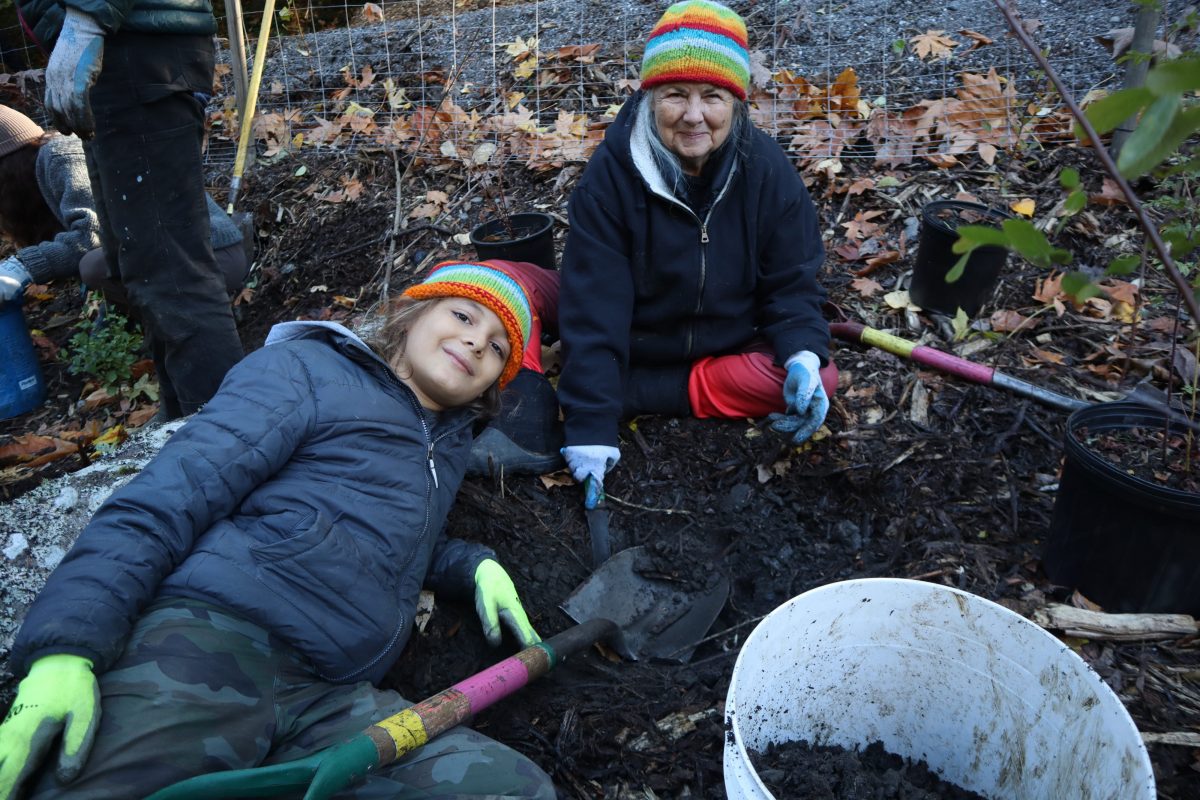 A child and adult wearing colorful hats and smiling while posing next to the plants they are planting outside.