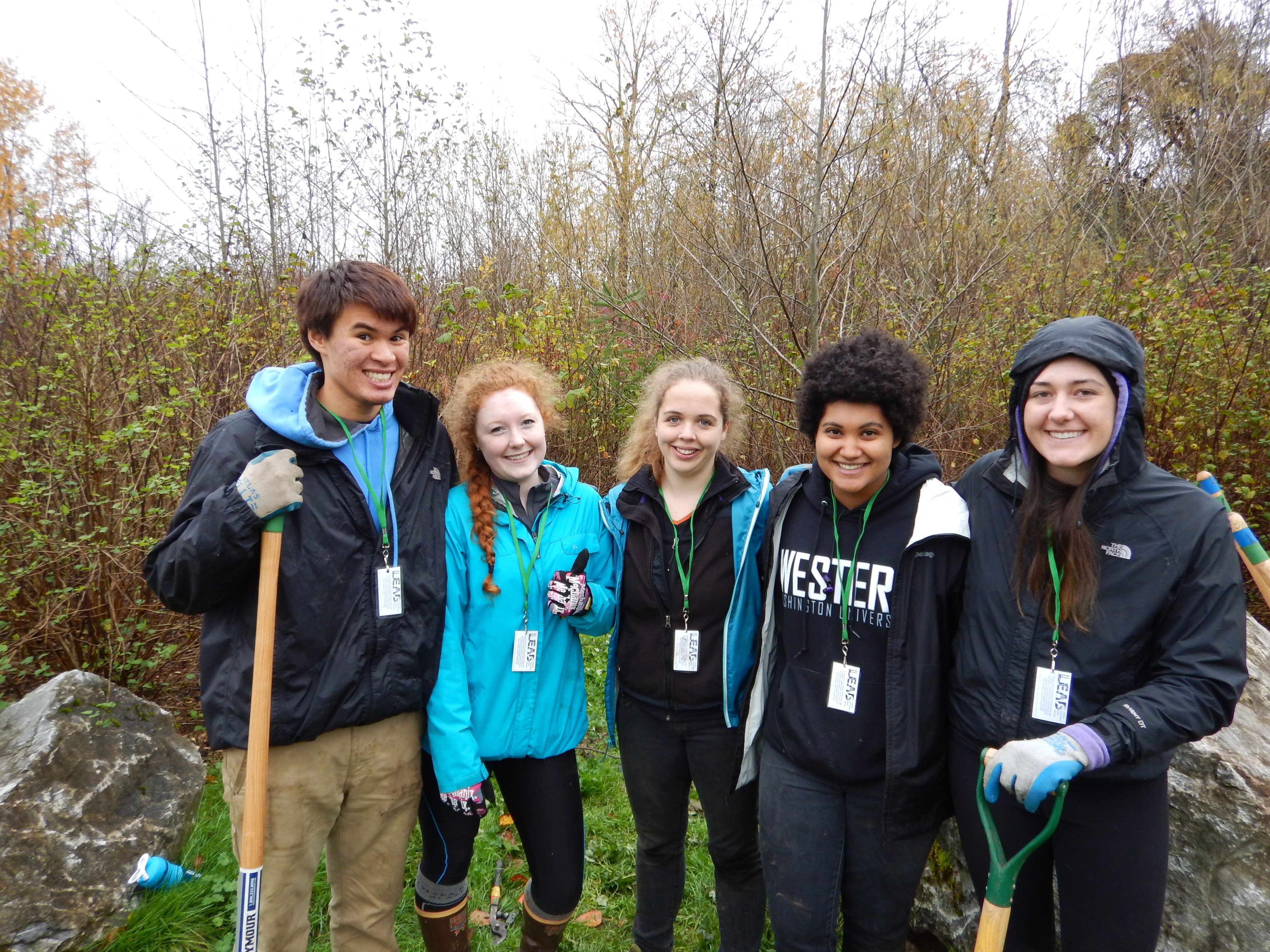 Five volunteers holding shovels and smiling together with trees in the background