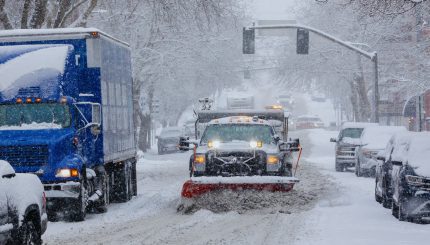 Truck with snow plow clearing snow on urban street with cars parked and driving nearby