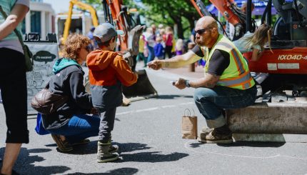 Worker wearing safety vest sitting on curb handing a kid a card with work trucks and people in the background