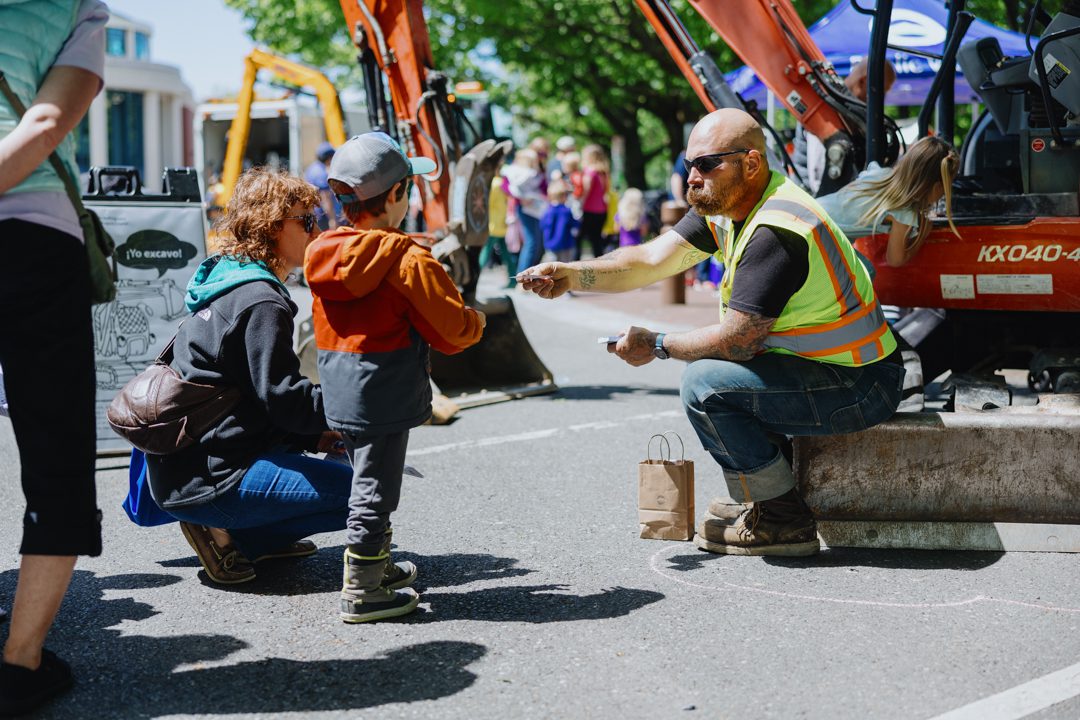 Worker wearing safety vest sitting on curb handing a kid a card with work trucks and people in the background
