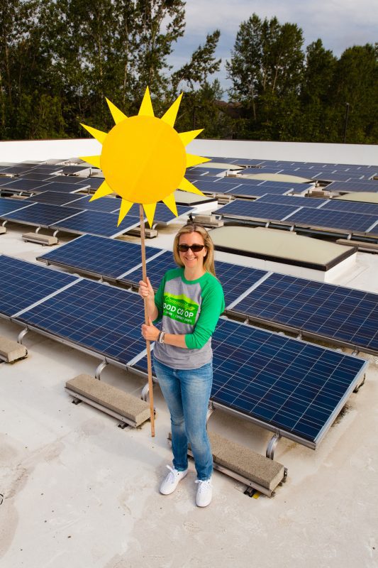 Person standing on roof of a building next to solar panel array holding a cut-out sun. 