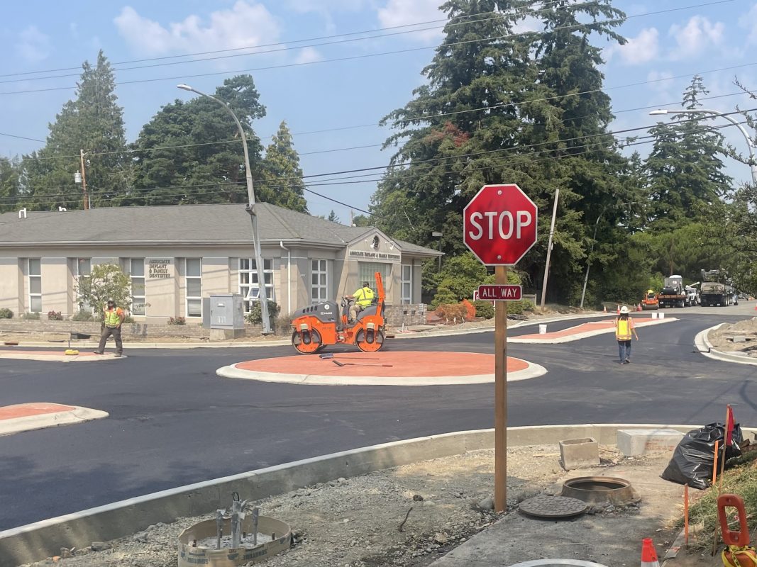 Newly paved intersection with construction workers working and round median in the center of the intersection.