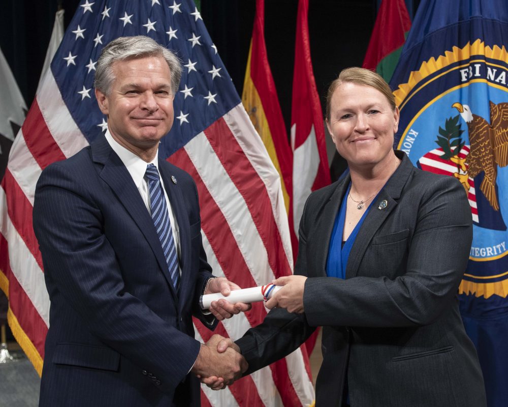 Two individuals in business suits smiling while shaking hands and passing rolled certificate with US flag in background