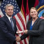 Two individuals in business suits smiling while shaking hands and passing rolled certificate with US flag in background