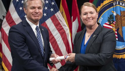 Two individuals in business suits smiling while shaking hands and passing rolled certificate with US flag in background