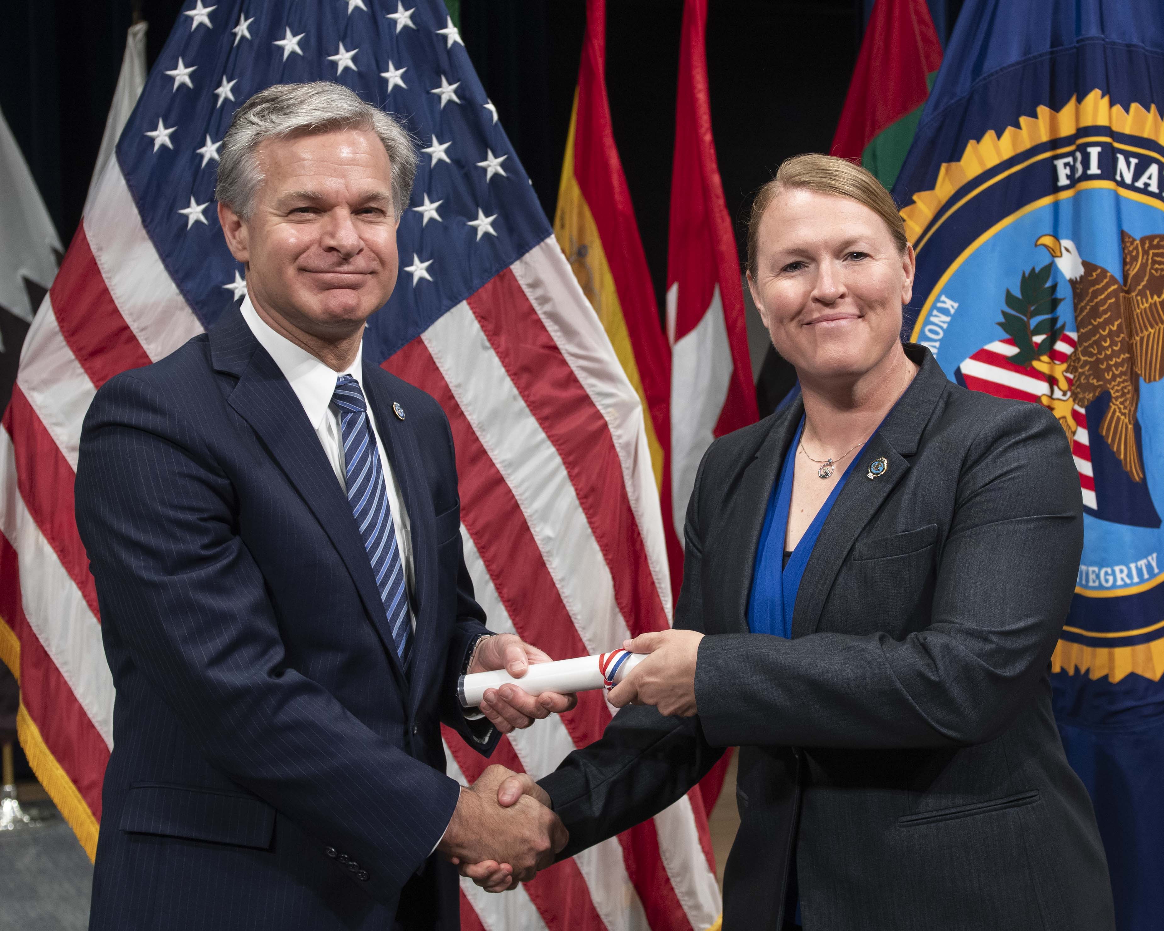 Two individuals in business suits smiling while shaking hands and passing rolled certificate with US flag in background