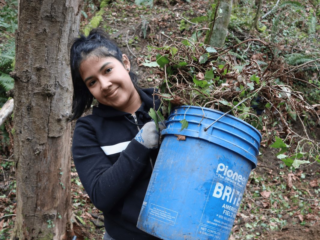 Young adult smiling, standing in forest, holding blue bucket overflowing with weeds