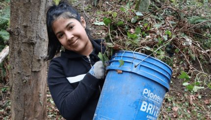 Young adult smiling, standing in forest, holding blue bucket overflowing with weeds