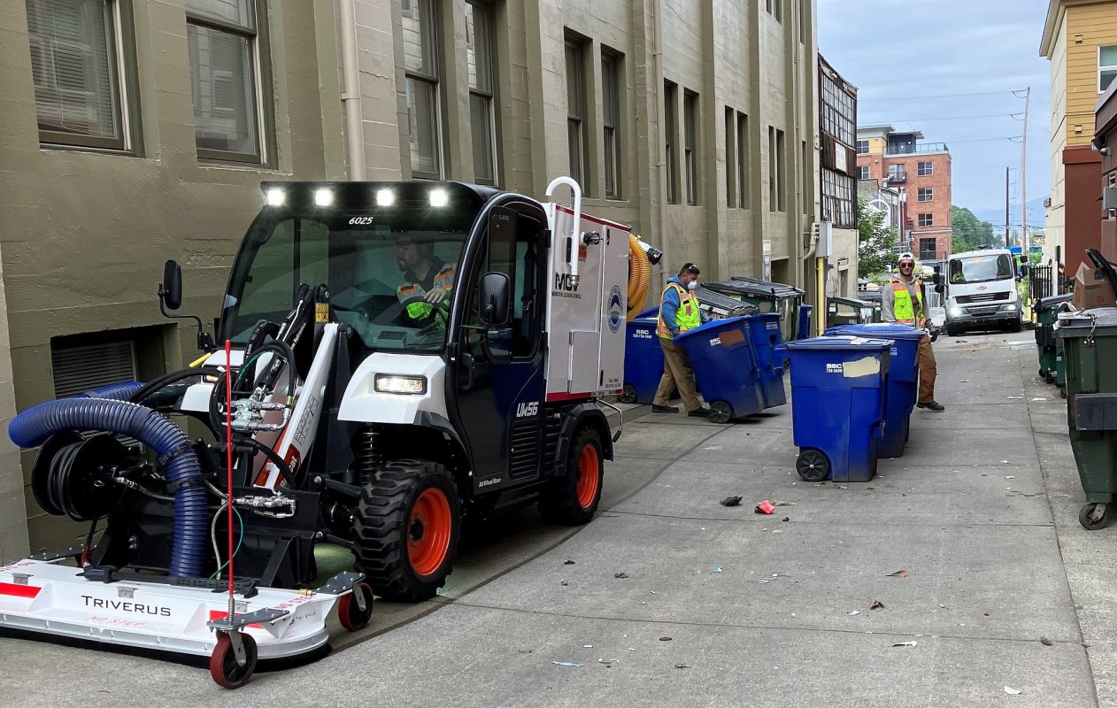 Workers in alleyway moving garbage and recycling bins while an operator drives a small sweeping device that says MCV on the side. 