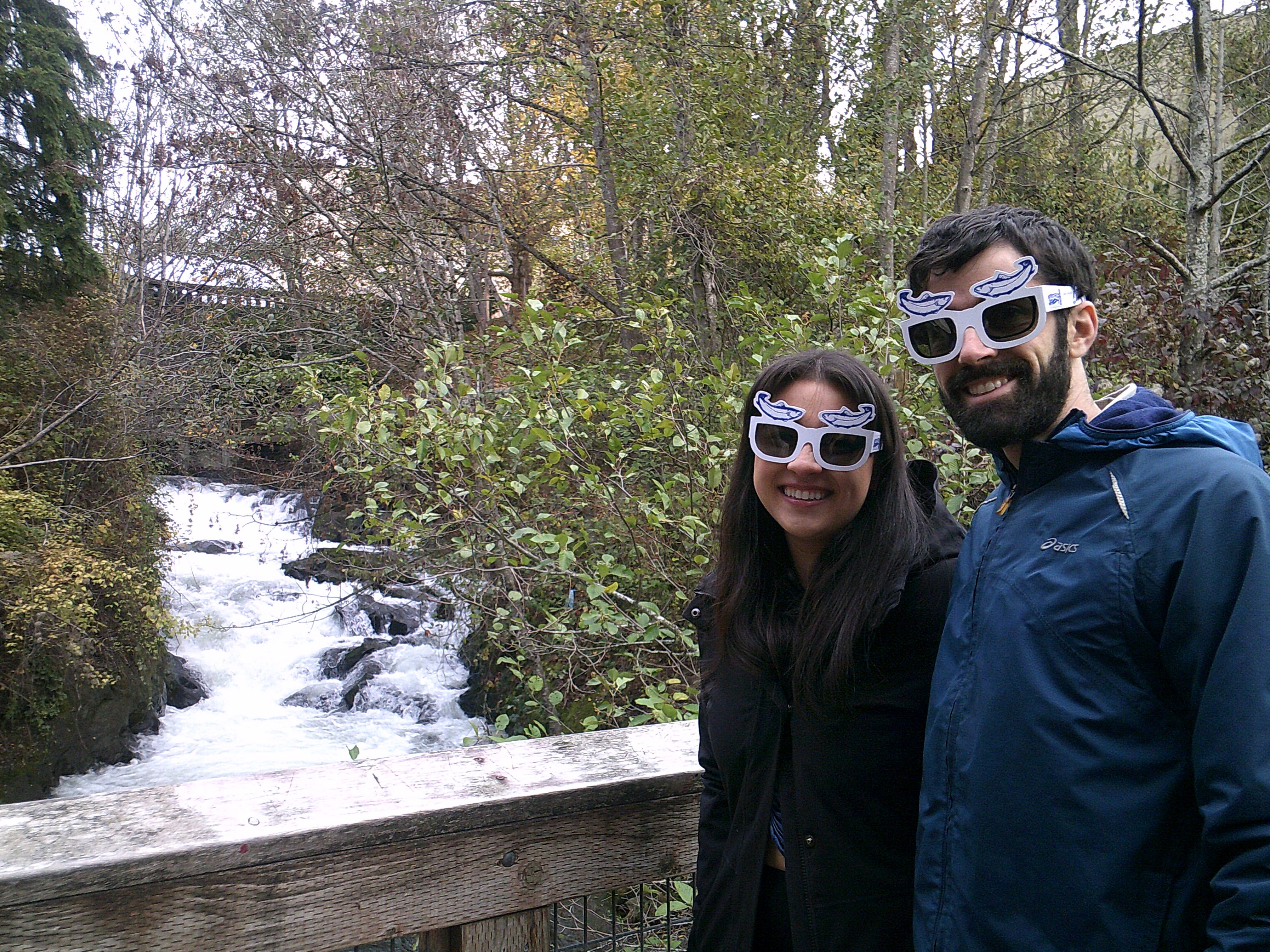 Two people standing on a bridge smiling wearing paper sunglasses with salmon on them. Creek flowing in the background.