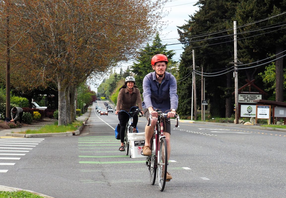 Two bike riders with helmets riding in a bike lane on a two-lane road through suburban area.