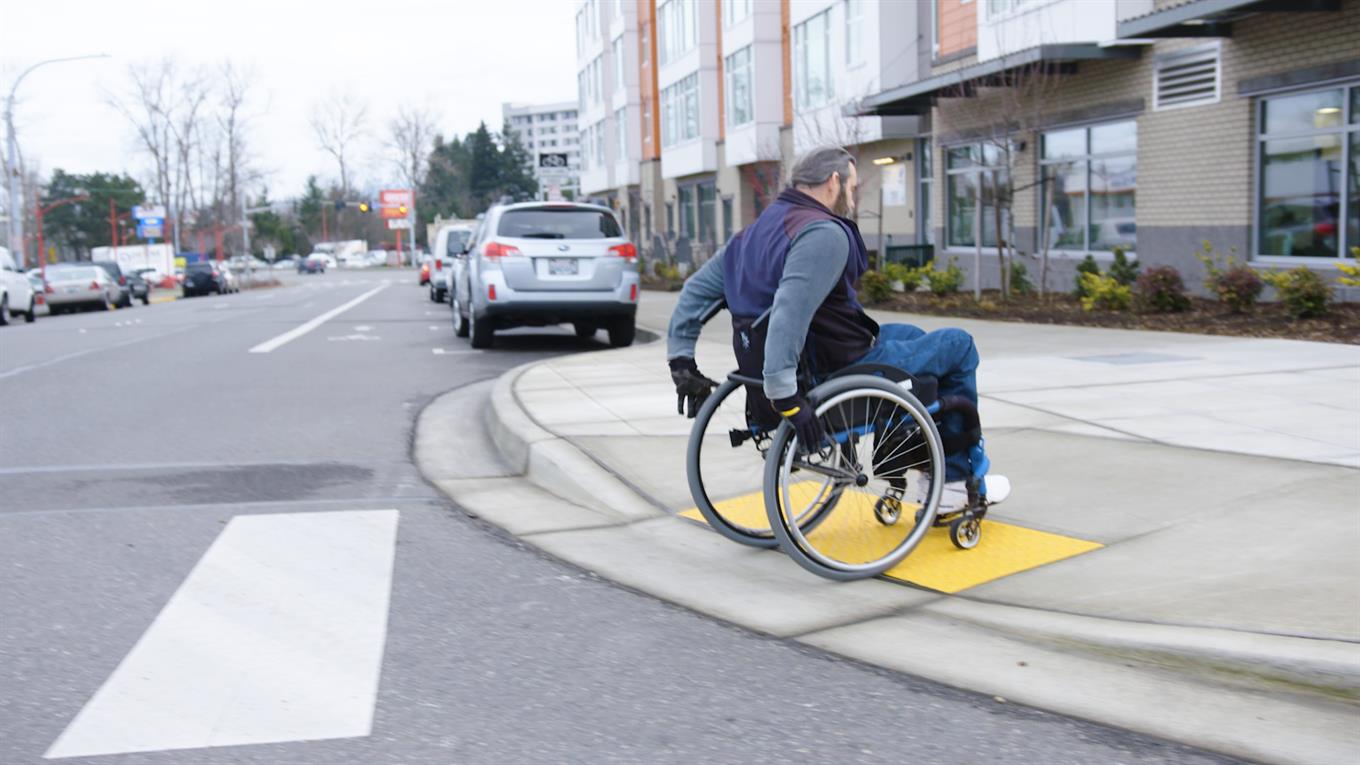 Individual in wheelchair wheeling themselves from crosswalk to sidewalk ramp