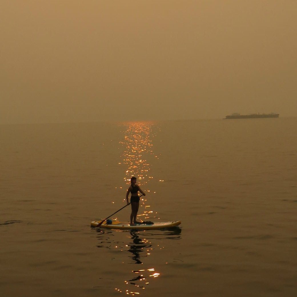 Person standing on a paddleboard in water with sun shining through a smoky sky.