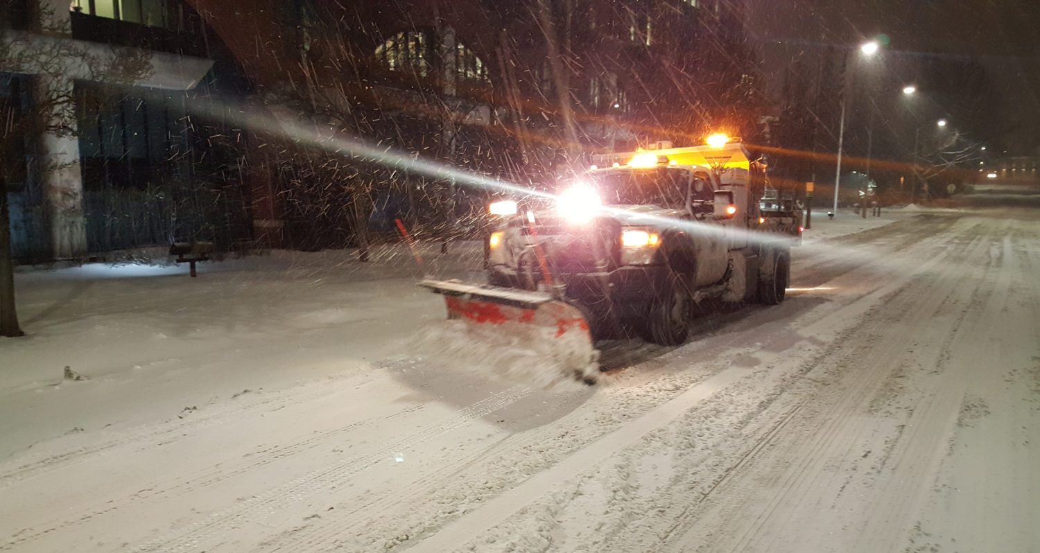 snow plow with head lights driving on snowy street in the dark