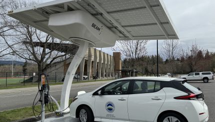 Small passanger vehicle plugged into charger with large solar panel over head.