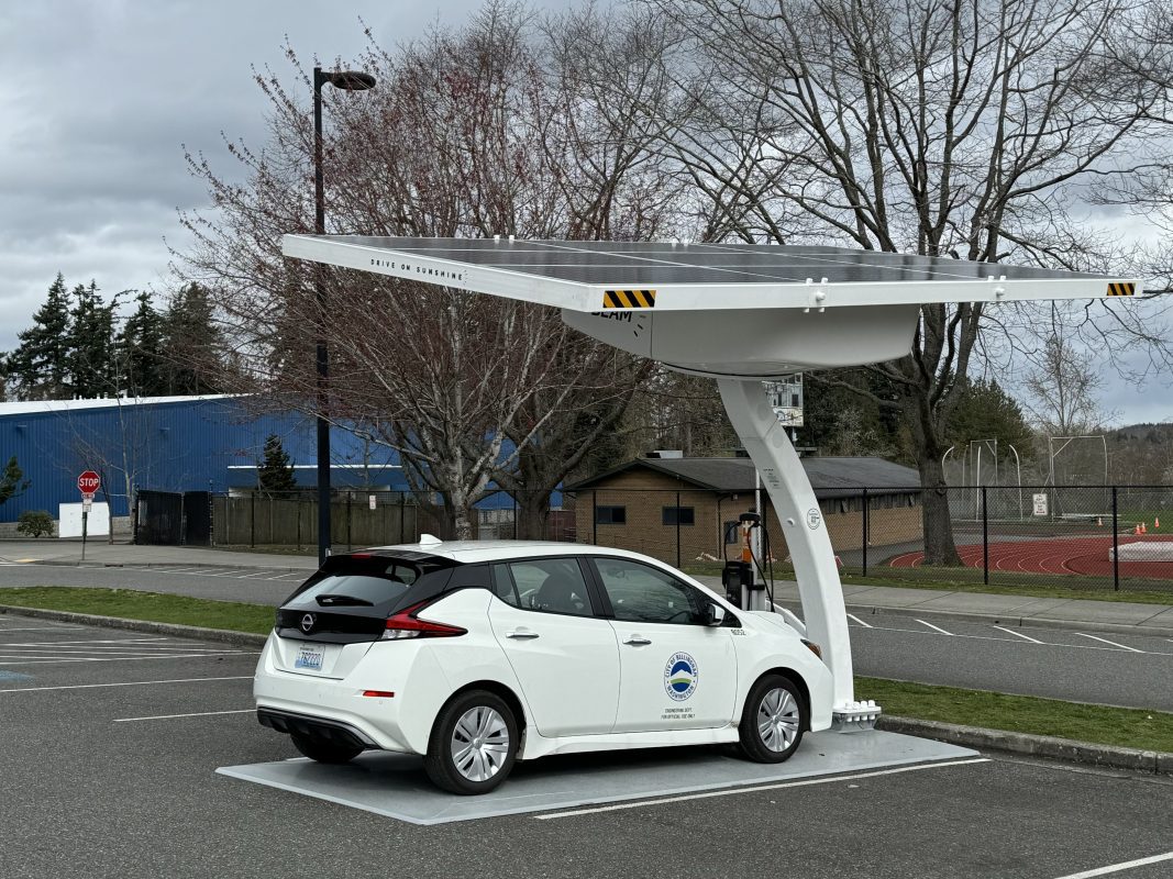 White electric Leaf vehicle plugged into a charging station with a very large solar panel above it