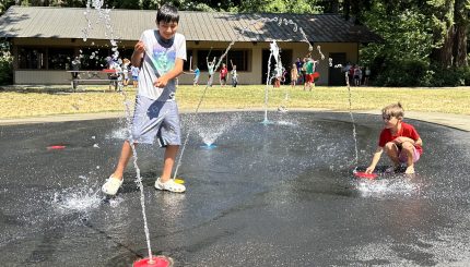 two children playing in water that is being sprayed from the ground in a park with many kids playing on field in the background