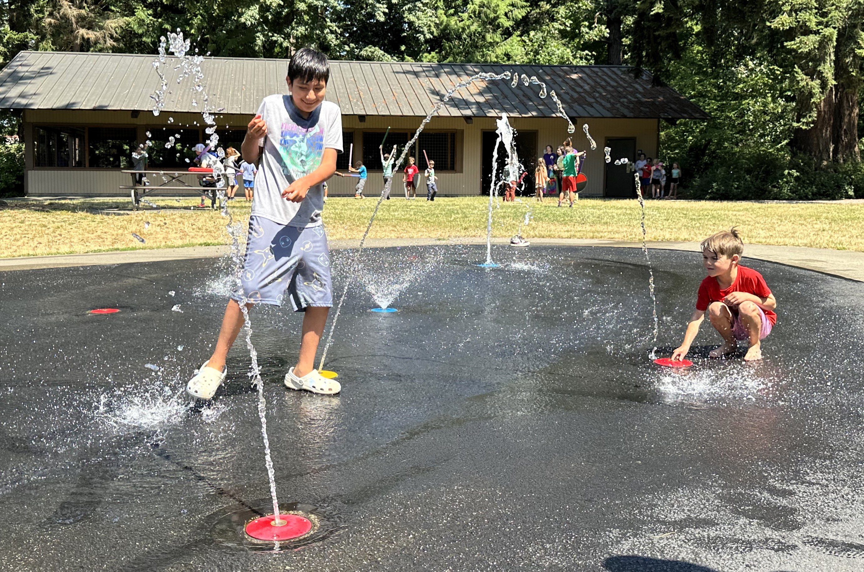 two children playing in water that is being sprayed from the ground in a park with many kids playing on field in the background