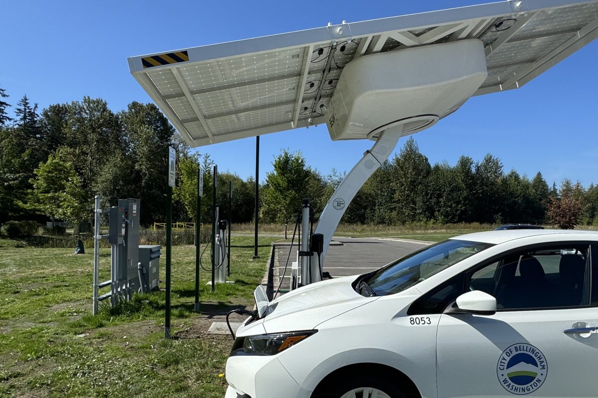 Electric vehicle charger with big solar panel array above it at a park. A city vehicle is plugged into one of the chargers.