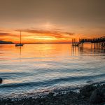 Sunsetting over marine waters with sailboat anchored in distance and large dock on the side