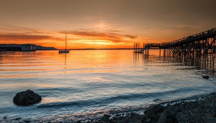 Sunsetting over marine waters with sailboat anchored in distance and large dock on the side