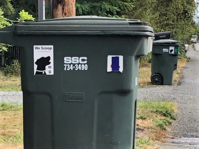 View of street with garbage cans lined up near the road. Garbage cans each have a 4 by 6 inch sticker that says We Scoop and the black silhouette of a dog.