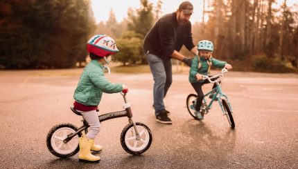 Two young girls with helmets riding bikes on pavement with man helping one on a peddle bike