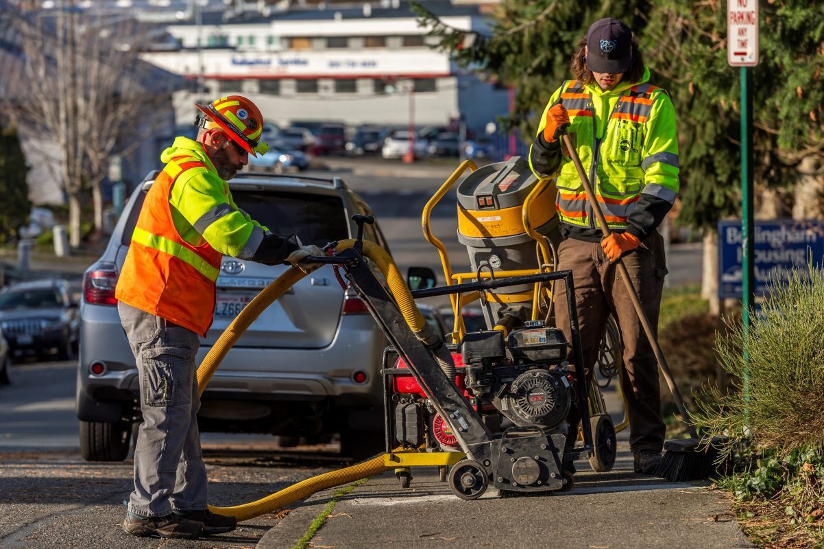 Two City of Bellingham workers in neon yellow and orange clothing operating machinery on a sidewalk.
